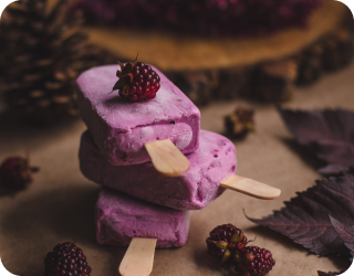 Três paletas empilhadas com uma amora em cima e decorações com pinhas, folhas e amoras na mesa